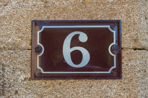 A traditional dark brown enamel house number 6 plaque with white numerals mounted on a rough-textured stone wall in a French village