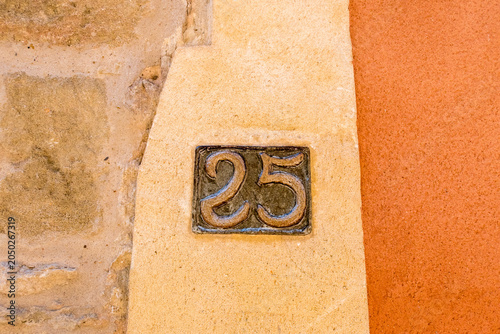A small, ornate bronze number 25 plaque set into a cream rendered pillar beside a vivid orange wall in a French village