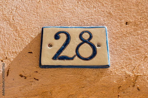 A ceramic house number 28 plaque mounted on a rustic terracotta orange rendered wall in a French village
