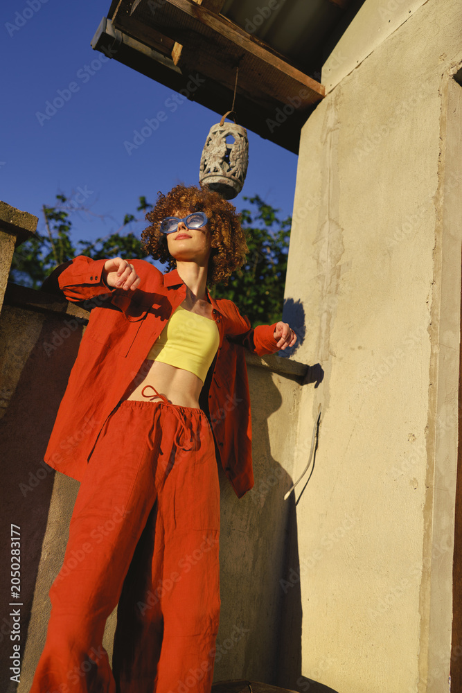 Obraz premium A woman in bold red-orange outfit and yellow top poses outdoors, leaning against a sunlit wall with a hanging lantern above, a lifestyle portrait of confidence and fashion