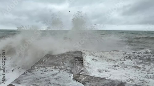 Ocean waves continuously crash against a concrete pier, with stormy clouds looming over the sea in a coastal landscape