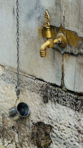 Vertical perspective view of a vintage brass faucet and hanging metal water jug at the ablution fountain of Eyup Sultan Mosque in Istanbul, Turkey. Ancient Ottoman architecture design details.