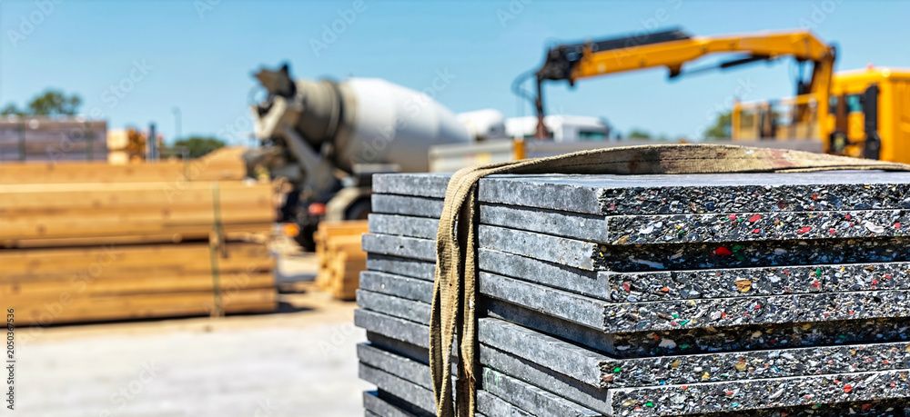 Naklejka premium Stack of gray recycled plastic panels with speckled cross sections. Sustainable construction materials at a building site with blurred concrete mixer in background