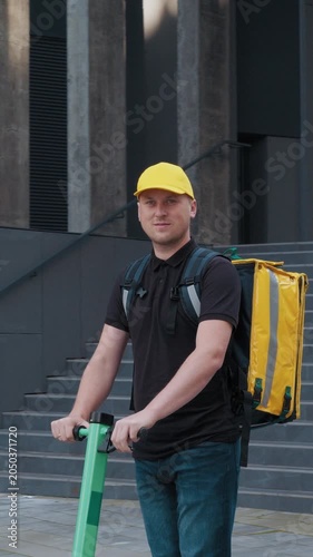 A delivery man wearing a yellow cap and carrying a backpack walks down the street, vertical video.