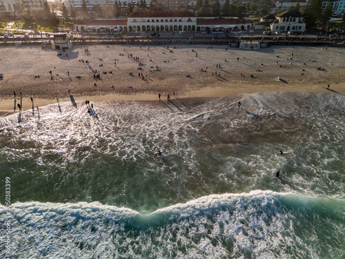 Bondi Beach aerial view crowded with swimmers, surfers and waves breaking in golden hour. Key buildings, pavilion and promenade can be seen in background.