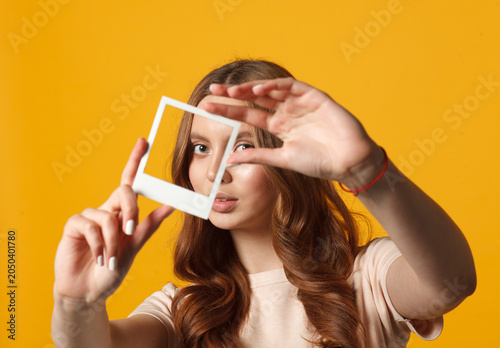 Young woman holding empty frame in front of her face against yellow background. Creative visual concept.