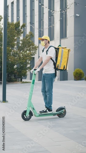 A delivery person wearing a mask and carrying a food delivery bag rides an electric scooter on a city street. Vertical video.