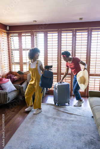 Diverse women packing backpack, pulling silver suitcase, holding straw hat and smartphone at home