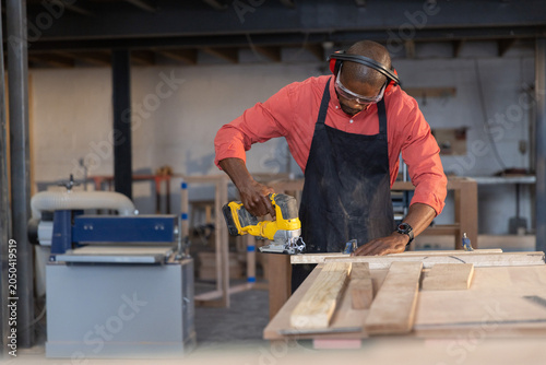 Woodworker is cutting wooden plank with yellow cordless jigsaw on workbench in carpentry workshop