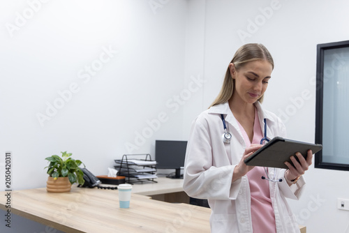 Adult clinician in lab coat and scrubs at clinic desk using tablet and stethoscope, copy space