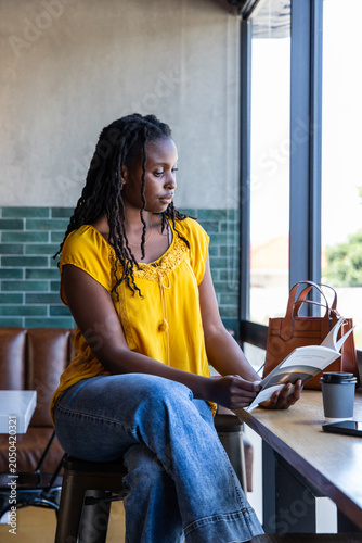 Adult African American woman in yellow blouse sitting at cafe counter reading book with coffee cup