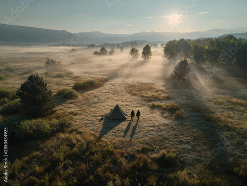Serene landscape featuring two hikers standing next to tent in misty meadow at sunrise. soft light creates tranquil atmosphere, highlighting beauty of nature