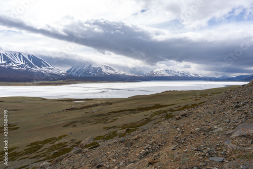 Frozen Alpine Lake with Melting Ice, Larch Forest and Snow-Capped Mountain Range Under Dramatic Clouds, Mongolian Altai