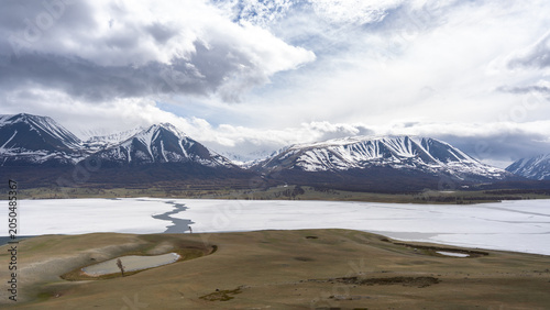 Frozen Alpine Lake with Melting Ice, Larch Forest and Snow-Capped Mountain Range Under Dramatic Clouds, Mongolian Altai