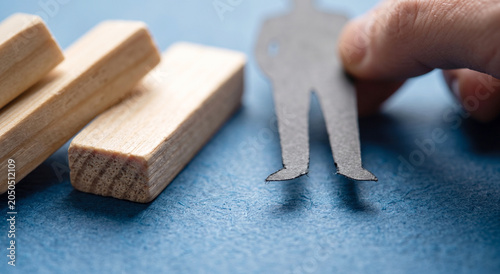 Hand holding a black paper cutout figure next to fallen wooden domino blocks on a blue desk background.