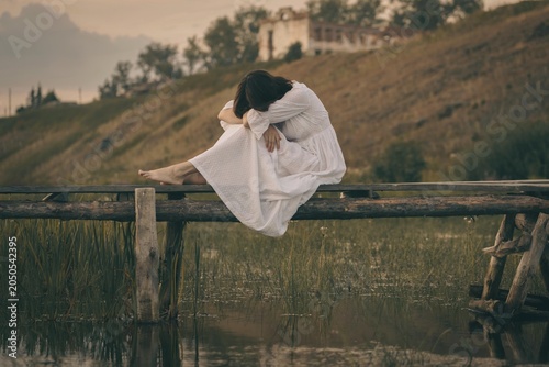 Authentic barefooted young woman in vintage white dress sits curled up on old wooden small bridge over a calm water in summer. Atmosphere of pagan witchcraft, earth magic and medieval