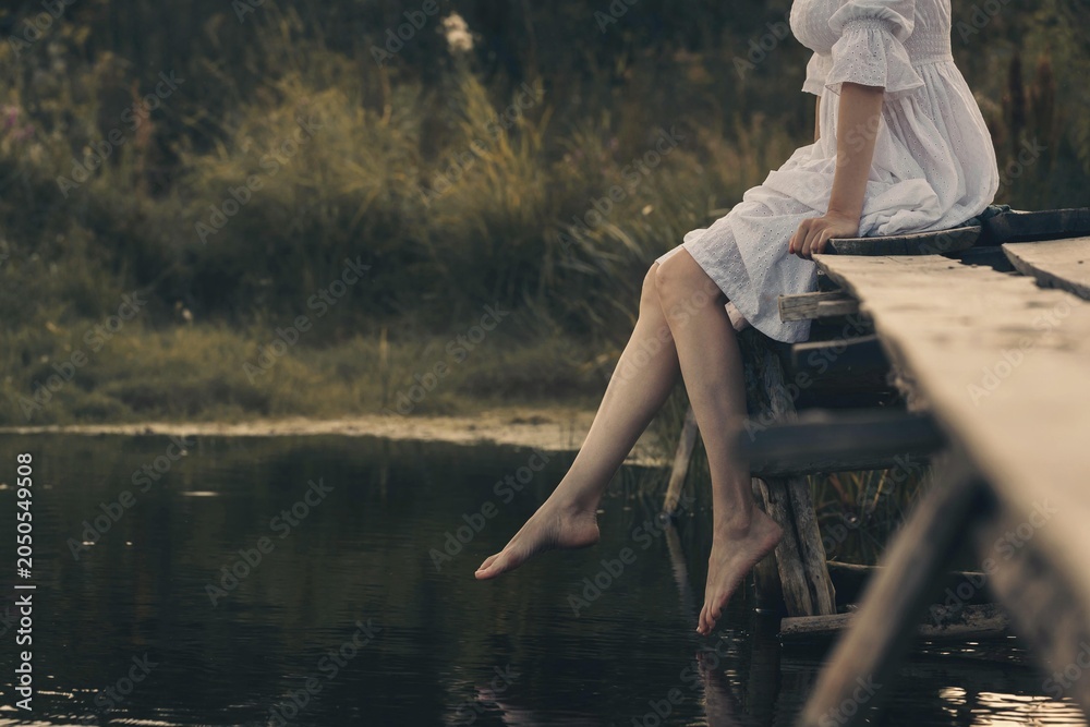 Naklejka premium Atmospheric scene of faceless young woman in white vintage dress sitting and swinging her bare legs on old wooden pier over dark wild pond in summer