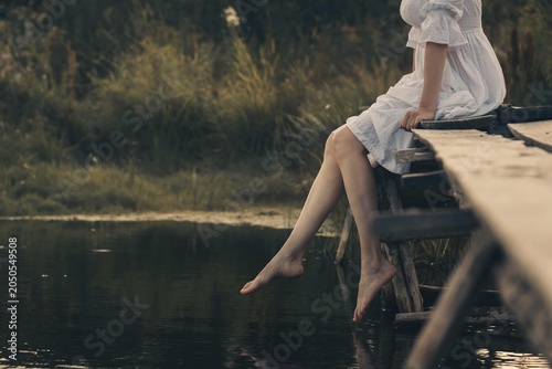 Atmospheric scene of faceless young woman in white vintage dress sitting and swinging her bare legs on old wooden pier over dark wild pond in summer