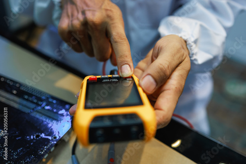 Obraz na plátně Close-up of hands checking a yellow handheld electronic meter beside a laptop, showing device testing, repair, or technical inspection in a modern workspace