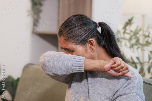 Middle aged woman coughing or sneezing into her elbow while sitting on a sofa at home, showing cold, flu, allergy or hygiene care in a cozy indoor setting.