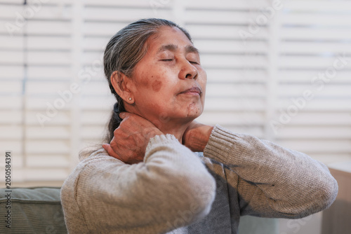 Senior woman at home holding her neck with eyes closed, showing pain, stiffness, stress or fatigue. Warm indoor portrait with soft light and calm background.
