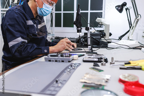 Quadro em tela Technician in safety mask and face shield repairs electronic parts at a workbench with tools, microscope and computer in a clean workshop
