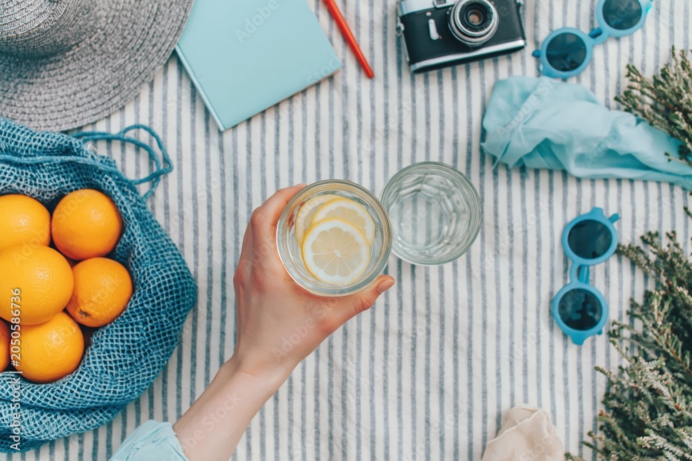 Fototapeta premium Picnic scene with hand holding refreshing lemonade glass, summer fruit, oranges, camera