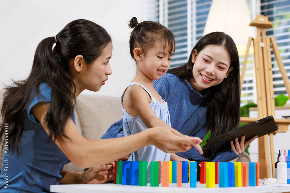 Obraz premium Two women and a young girl play with colorful blocks while looking at a tablet in a bright living room, showing family learning, bonding, and fun at home.