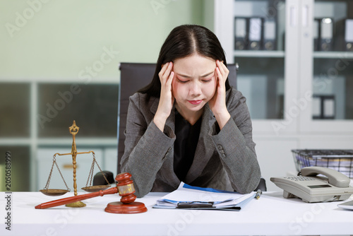 Stressed female lawyer sits at an office desk holding her head, with legal papers, gavel and scales nearby, showing work pressure, headache and office fatigue.