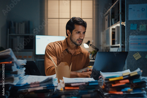 Young Latin businessman eating takeout instant noodles and reviewing document on laptop at desk in dark night office. Overtime working late, overworked deadline, fast food, corporate problem concept.