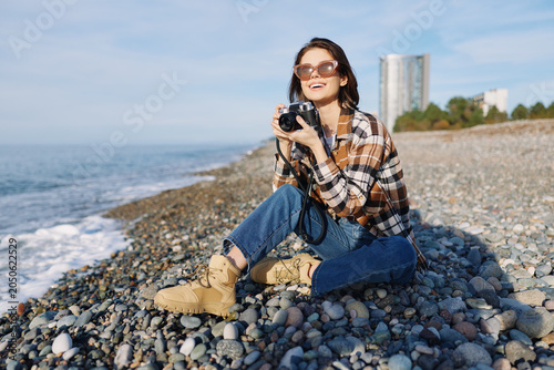 Tableau sur toile Woman photographer on a pebble beach with a camera, wearing a plaid shirt, denim jeans and boots, enjoying sunny coastal scenery and open sea view at the shore