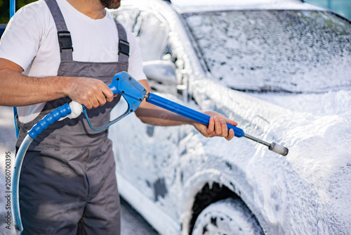 Schilderij op canvas A person in overalls uses a high-pressure washer to spray foam onto a car during