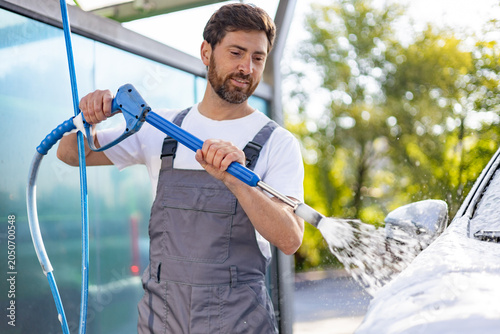Quadro su tela A caucasian man in overalls uses a high-pressure washer to clean a car covered i