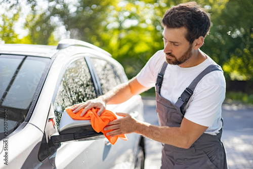 Billede på lærred A caucasian man in overalls meticulously wipes down a car's side mirror with an