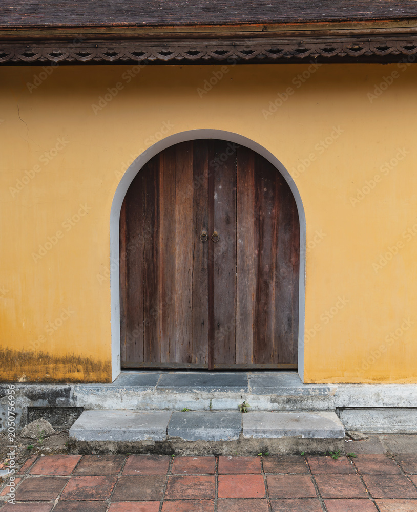 Fototapeta premium Traditional arched wooden door set in a vibrant yellow wall with ornate roof details at the Imperial City, Hue, Vietnam