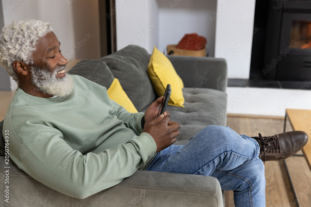 Obraz premium Senior African American man reclining on sofa at home using smartphone in sweatshirt, copy space