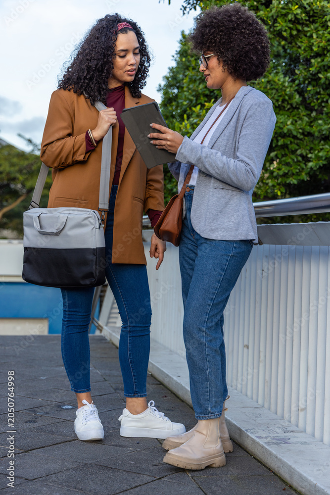 Fototapeta premium Diverse female colleagues leaning in, discussing tablet on walkway by metal railing wearing blazers
