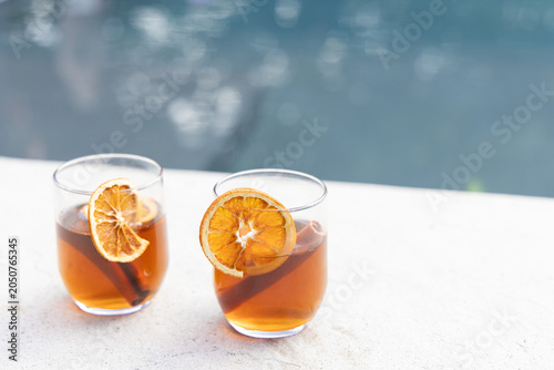 Two glasses sitting on pool ledge with amber drinks, orange wheels, cinnamon sticks, copy space