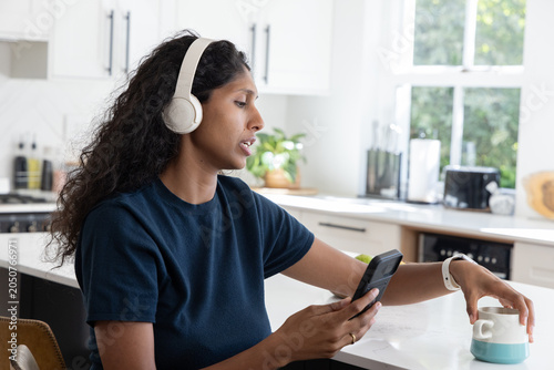 African American woman sitting in kitchen wearing navy tee, headphones, holding phone, reaching mug