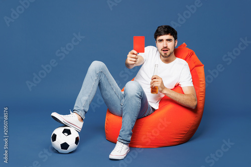 Fotografija Full body sad upset young man fan wear white t-shirt cheer up football sport team hold in hand red card soccer ball watch tv live stream sit in bag chair drink beer isolated on plain blue background