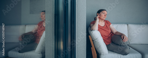 Fotografía Pensive middle-aged woman sits alone on a couch beside a glass door, gazing outw