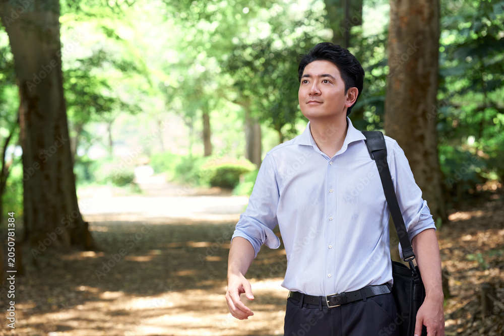 Fototapeta premium 緑豊かな公園を歩く30代の日本人男性会社員が鞄を肩にかけて爽やかな表情で遠くを見つめる様子