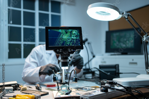 Fotografi Engineer in a lab repairing a circuit board under a digital microscope, with tools, screen and bright desk lamp in a modern electronics workspace