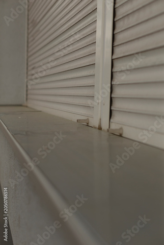 Close-up view of a closed white metal roller shutter mounted over a window sill. The image shows horizontal slats, metal frame, and a flat painted concrete or