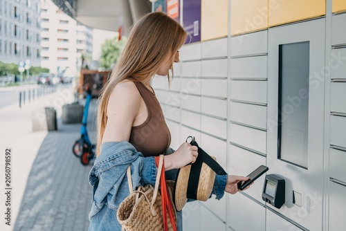Woman presents smartphone to automated parcel station scanner while holding bags and straw hat. Click and collect retail service, online shopping parcel pickup, consumer convenience economy lifestyle.