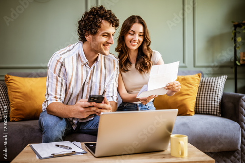 A woman holds a bill or statement while speaking with her partner on the sofa. Their are working together to understand expenses, manage finances, or plan upcoming payments.
