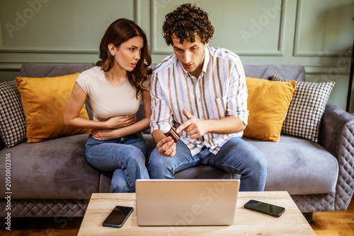 Adult couple sitting on bed in living room and having online doctor's appointment using laptop. Woman having pain in stomach. Remote consulting with doctor.