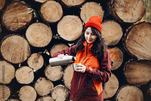Photos Smiling woman pours drink from metal thermos into cup while standing in front of stacked logs in forest