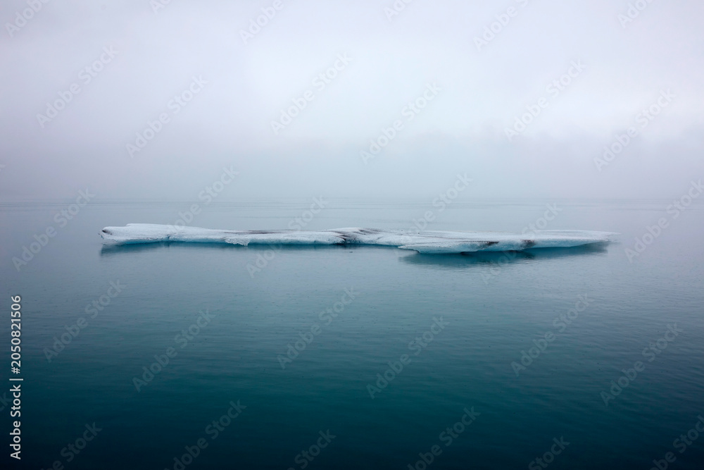 Naklejka premium Isolated minimalist ice floe floating in calm turquoise glacial lagoon with thick fog in horizontal Iceland view