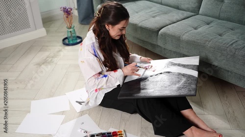 Focused young woman artist painting canvas at home. Beautiful creative female painter holding watercolor palette and drawing artwork on floor in living room. Hobby and art therapy concepts, talent